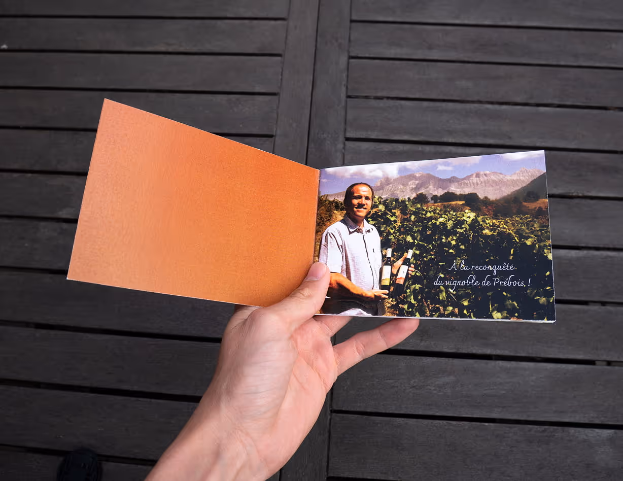 Hand holding an open brochure showing a man in a vineyard holding two wine bottles with mountains in the background and French text on the right page.