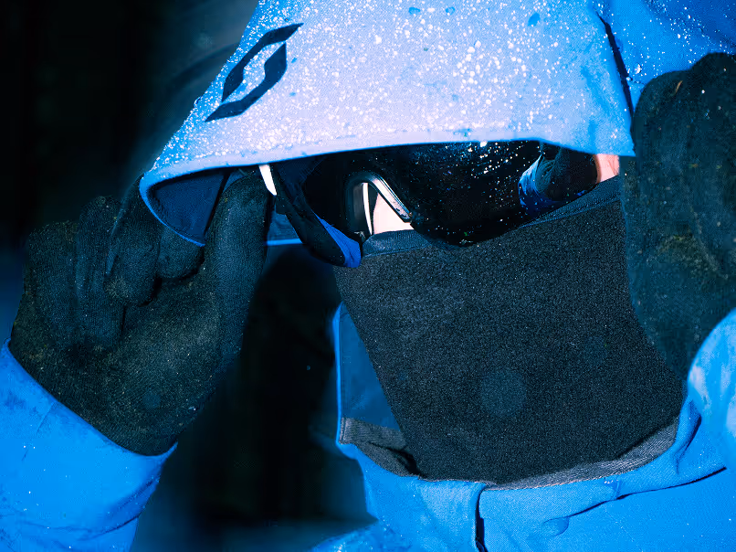 Close-up of a person wearing a blue waterproof hooded jacket, black gloves, dark goggles, and a black face covering in a snowy or rainy environment.
