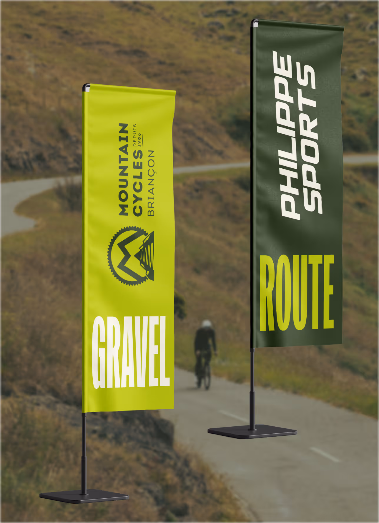 Deux oriflammes en bord de route de montagne, « Mountain Cycles Briançon – Gravel » et « Philippe Sports – Route », un cycliste au loin