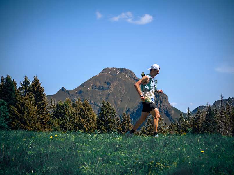 Coureur en casquette et short trottant sur une prairie herbeuse avec montagne et forêt en arrière-plan.